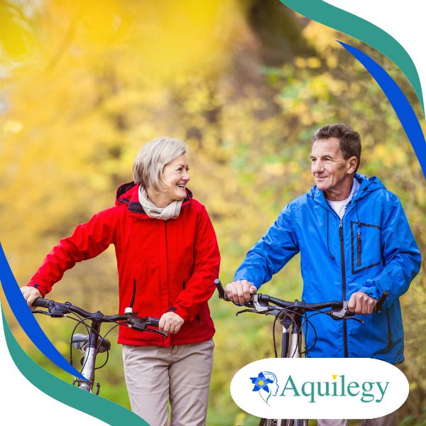 An older man and woman in red and blue jackets walk with bicycles on a tree-lined path. The Aquileg logo is at the corner.