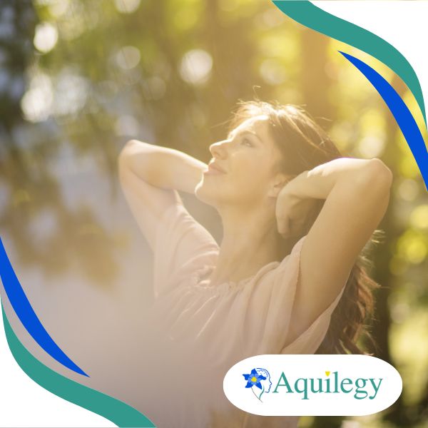 A woman is outdoors, looking up with her hands behind her head, surrounded by trees. The image has a soft, blurry background. A logo in the corner reads "Aquileg.