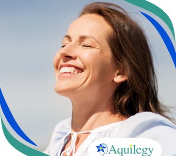 A woman with closed eyes and a broad smile stands outdoors in sunlight. The Aquilegy logo appears in the bottom right corner.