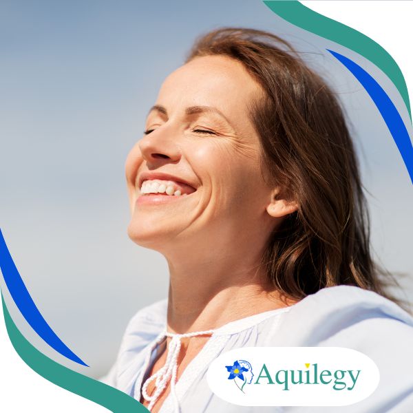 A woman with closed eyes and a broad smile stands outdoors in sunlight. The Aquilegy logo appears in the bottom right corner.
