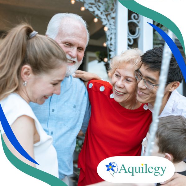 Four people of different ages stand close together, smiling and embracing outdoors. The Aquilegy logo is visible in the bottom right corner.