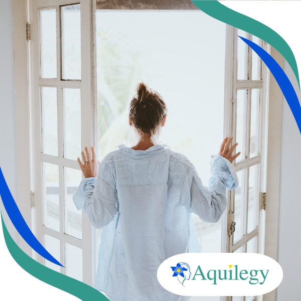 A person in a light shirt stands with their back to the camera, opening glass doors to a bright outdoor area. Aquilegy logo is displayed in the bottom right corner.