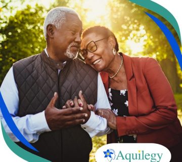 An older couple smiles and stands close together outdoors, with the woman leaning her head on the man's shoulder. The Aquilegy logo appears in the bottom right corner.