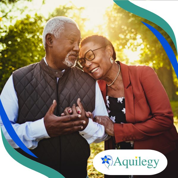 An older couple smiles and stands close together outdoors, with the woman leaning her head on the man's shoulder. The Aquilegy logo appears in the bottom right corner.